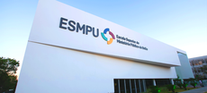 Foto do prédio da ESMPU, que tem a cor branca. Na fachada superior, está a logomarca da instituição, composta pela sigla ESMPU e um cata-vento estilizado nas cores amarela, azul claro, azul escuro e vermelho.